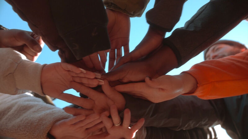 The image shows a group of people with their hands stacked on top of each other, forming a circle. The hands are of various skin tones, suggesting a diverse group. The composition is from a low angle, looking up at the hands against a blue sky. The overall impression is one of unity, teamwork, and collaboration.